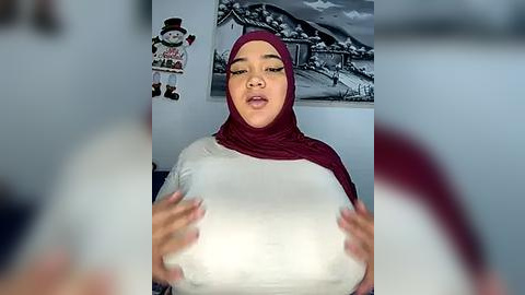 Media: Video of a plus-sized woman with large breasts, wearing a maroon hijab and a white top, holding her breasts. Background features a festive snowman decoration and a black and white photo of a snowy landscape.