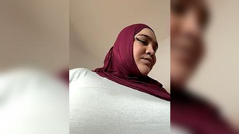 Media: Video of a smiling, dark-skinned woman with a maroon hijab and white top, standing in a beige room.