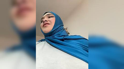 Media: A video of a woman in a blue hijab and white top, looking down with a slight smile. The background is out of focus, creating a blurred effect.