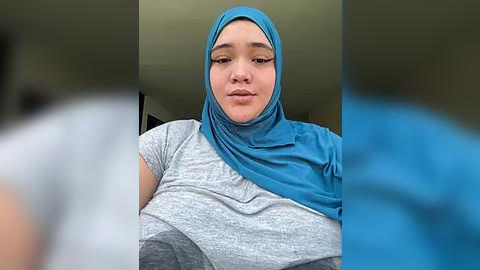 Media: Video of a young woman with light skin, wearing a blue hijab and gray t-shirt, sitting indoors with blurred background.