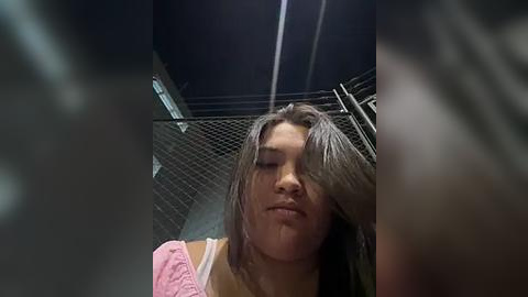 Media: Video of a young woman with long, wet hair, wearing a pink top, standing in a dimly lit, industrial bathroom with metal walls and a chain-link ceiling.