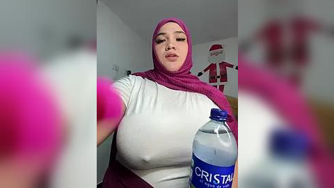 Media: Video of a young woman with a light complexion, wearing a magenta hijab and white top, holding a blue \"Cristal\" water bottle. The background features a white wall and a red and white striped shirt hanging.