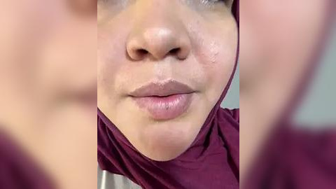 Media: Close-up video of a woman's face with light brown skin and full lips, wearing a maroon hijab. The background is out of focus, highlighting her facial features.