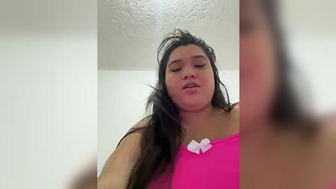 Media: Video of a young Latina woman with long, dark hair, wearing a bright pink tank top with a white bow, standing against a white wall with a textured ceiling. Her eyes are closed and she has a slight smile.