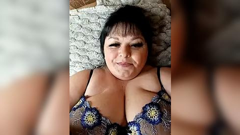 Media: A video of a middle-aged woman with a medium complexion and black hair, lying on a textured beige rug. She wears a blue and yellow floral bra, with visible cleavage. The background is blurred.