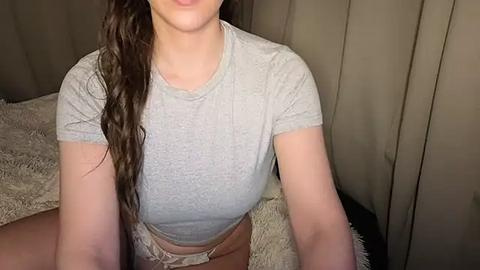 Media: A video of a light-skinned woman with long brown hair, wearing a tight gray t-shirt and beige shorts, sitting on a beige, textured bedspread, with a neutral-colored curtain backdrop.