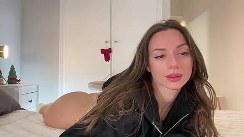 Media: Video of a young woman with long brown hair, wearing a black jacket, lying on her stomach on a white bed. Background includes a white wardrobe with red bows, a nightstand with a small Christmas tree, and a soft, warm lighting ambiance.