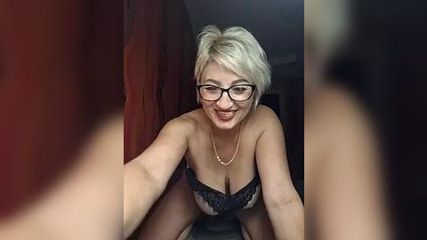 Media: A video of a smiling, light-skinned woman with short, platinum blonde hair and glasses, wearing a black lace bra, leaning forward in a dimly lit room with blurred, out-of-focus figures in the foreground.