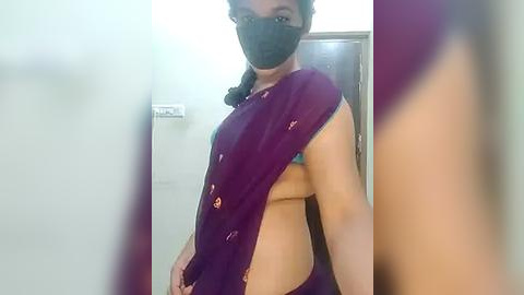 Media: Video of a South Asian woman with medium skin tone, wearing a purple sari with a black face mask, revealing her bare back and small breasts. Background features a plain white wall and a door.