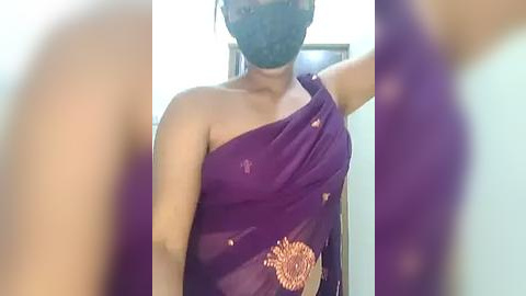 Media: Video of a woman wearing a purple saree with golden embroidery, black mask, and black hair, standing indoors with blurred background.