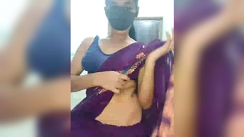 Media: A video of a woman in a blue top, black mask, and purple saree, holding a colorful object in a blurred indoor setting.