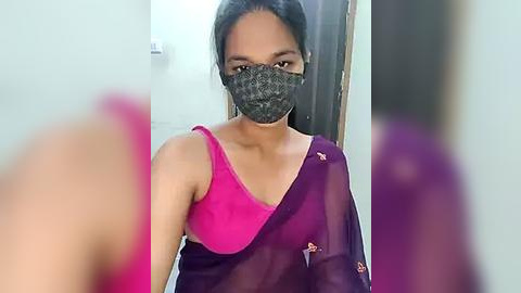 Media: Video of a woman with medium skin tone, wearing a bright pink bra and a sheer purple saree with floral embroidery, black face mask, and long dark hair, indoors, blurred background.