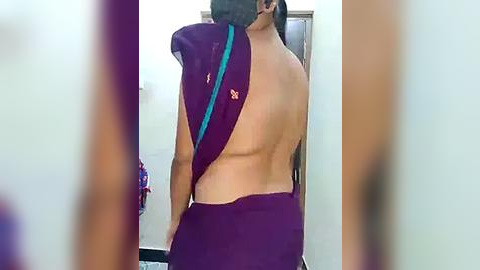 Media: Video of a woman with light brown skin, long black hair, and wearing a purple sari with a blue shoulder strap, captured from behind in a room with white walls and a mirror.