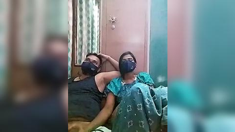 Media: Video of two people wearing masks, lying in bed, covered in blankets, with blurred background.