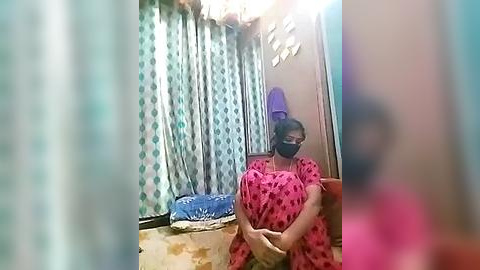 Media: Video of a woman in a pink dress with a heart pattern, wearing a face mask, standing in a cramped, dimly lit room with blue and white patterned curtains and a purple hat on a chair.