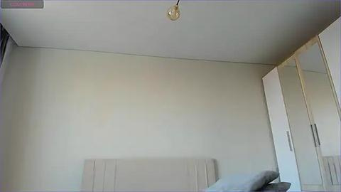 Media: A video of a minimalist, modern bedroom with a beige wall, a small, round light fixture, a bed with a padded headboard, and a mirrored wardrobe.