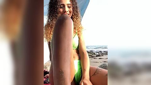 Media: Video of a young woman with curly, light brown hair and a light brown complexion, wearing a green bikini, sitting on a beach with her legs spread, sand on her thighs, and a blurred man in the foreground.