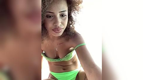 Media: Video of a young Black woman with curly hair, wearing green lingerie, capturing a moment of vulnerability. She has a slender physique, medium-sized breasts, and a subtle, natural makeup look.