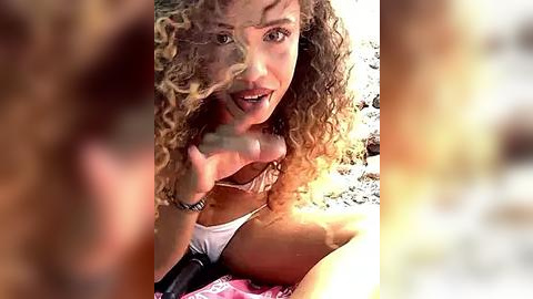 Media: Video of a woman with curly, blonde hair, smiling, holding a microphone, wearing a white bikini top. Blurred background with natural lighting.