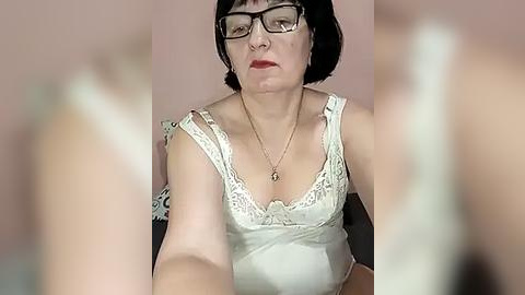 Media: A video of a middle-aged woman with short black hair, glasses, and a white lace camisole, sitting indoors against a pale pink wall. Her facial expression is neutral.