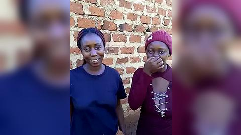 Media: Video of two smiling African women, one with a red headscarf, standing against a red-brick wall, with a blurred figure in the foreground.