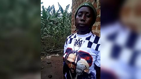 Media: A video of a young African boy with dark skin, wearing a white and black checkered shirt with a graphic of a man, standing against a brick wall and lush green foliage background.