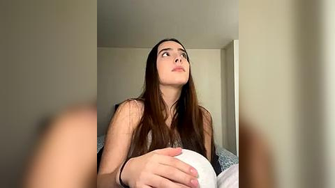 Media: Video of a young woman with long brown hair, light skin, and a contemplative expression, holding a white pillow, against a beige wall background.