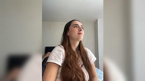 Media: A video of a young woman with long brown hair, wearing a white T-shirt, gazing upward in a minimalist, beige-walled room with a black TV and blue-patterned bedding.