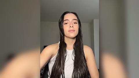 Media: Video of a young woman with long, wet, dark hair, wearing a white tank top, standing in a minimalist room with beige walls.