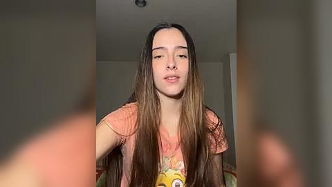 Media: Video of a young woman with long, straight brown hair, wearing an orange shirt with a cartoon character. She has a neutral expression, standing indoors with blurred, out-of-focus faces in the foreground.