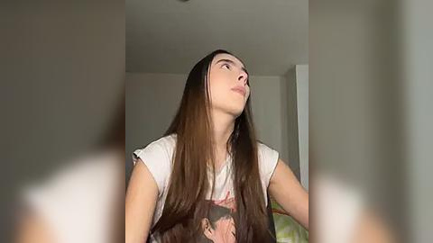 Media: Video of a young woman with long, straight brown hair, wearing a white T-shirt with a graphic, gazing upward with a thoughtful expression, in a dimly lit room.