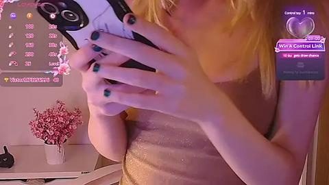 Media: A video of a woman with light skin, blonde hair, and green nail polish, wearing a shimmering gold dress, taking a selfie with a smartphone in a dimly lit room.