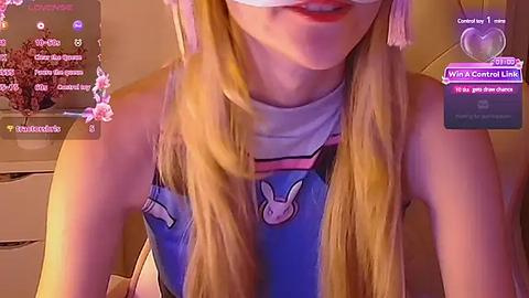 Media: Video of a young woman with long blonde hair, wearing a sleeveless blue shirt with bunny ears, in a cozy room.