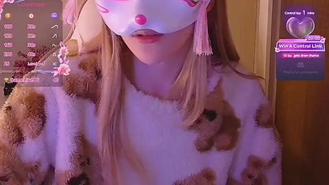 Media: Video of a light-skinned woman with long blonde hair, wearing a white mask and a plush, brown and white teddy bear sweater. Background includes a wooden door and live stream chat window with hearts and messages.