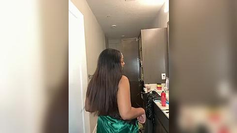 Media: A video of a woman with long, straight brown hair, wearing a green satin top, standing in a narrow hallway with beige walls and a closed door. The background features a messy kitchen counter with scattered items.