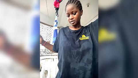 Media: Video of a young African woman with braided hair wearing a black T-shirt with yellow text, holding a red and blue object in a clean, well-lit room with white walls and a metal mesh screen.