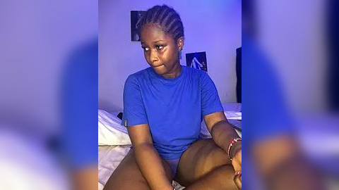 Media: Video of a young Black woman with braided hair, wearing a blue shirt, sitting on a bed in a dimly lit room, blurred background.