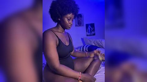 Media: Video of a dark-skinned woman with a natural afro, wearing a black tank top, sitting on a bed in a dimly lit bedroom with blue lighting.