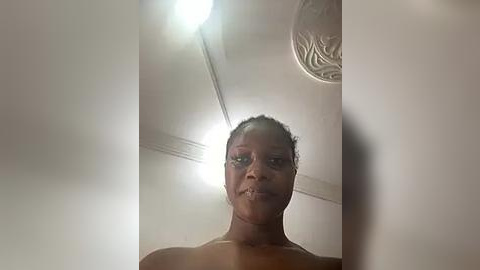 Media: Video of a topless Black woman with braided hair, wearing glasses, standing against a plain, off-white wall with a ceiling light and fan visible.