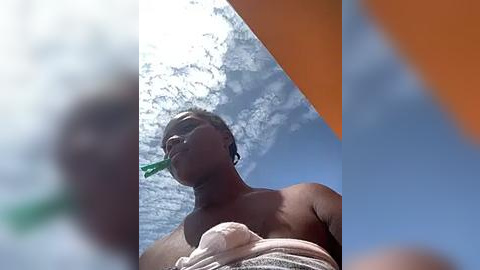 Media: Video of a shirtless Black man with short hair, wearing a white towel, eating a green leaf, against a bright blue sky with fluffy white clouds.