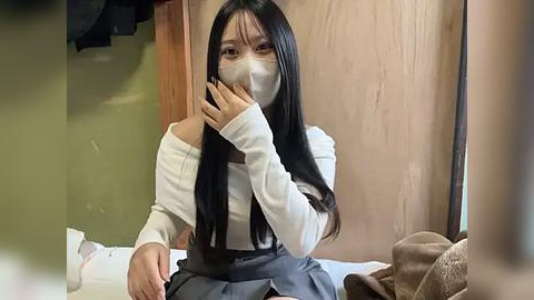 Media: Video of a young Asian woman with long black hair, wearing a white off-shoulder top and grey skirt, seated on a bed, covering her mouth with her hand, and wearing a face mask in a dimly lit room.