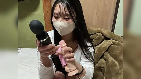 Media: Video of an Asian woman with long black hair, wearing a white mask, applying makeup with a black brush and a pink dildo, sitting on a bed with a beige blanket.