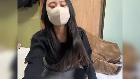 Media: Video of a woman with long, straight black hair, wearing a beige face mask, black shirt, and gray pants, sitting on a bed with a brown blanket and wooden door in the background.