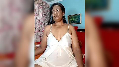 Media: Video of a middle-aged Black woman with medium skin tone, medium build, and medium-length black hair, wearing a white satin camisole, sitting on a bed with floral wallpaper and a red bedspread.