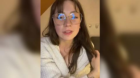 Media: Video of a young woman with long dark hair, wearing glasses and a light-colored knit cardigan, sitting indoors with a blurred background.