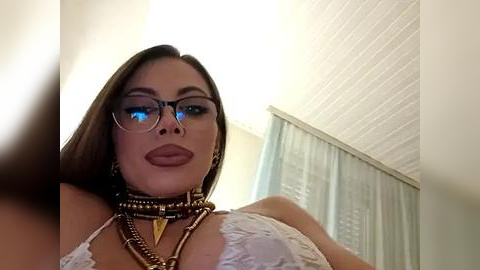 Media: A video of a light-skinned woman with long, dark hair, wearing black-rimmed glasses, a white lace bralette, and multiple gold necklaces. She stands in a brightly lit room with white walls and a light-colored curtain.