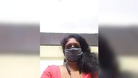 Media: A video of a Black woman with curly hair, wearing a gray mask and red shirt, standing in a bright, white-walled room with a blurred, gray object in the foreground.