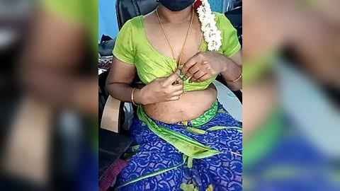 Media: Video of a South Asian woman with medium brown skin, wearing a green blouse and blue sari, seated in a car. She has a white flower in her hair and a mask covering her nose and mouth.