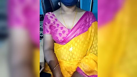 Media: Video of a woman in a bright yellow sari with pink borders, wearing a blue face mask and gold necklace, seated in a car.