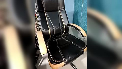 Media: A video of a black leather office chair with wooden armrests, positioned against a teal wall. The chair has a sleek, modern design with visible stitching and a cushioned seat. The background is slightly blurred.